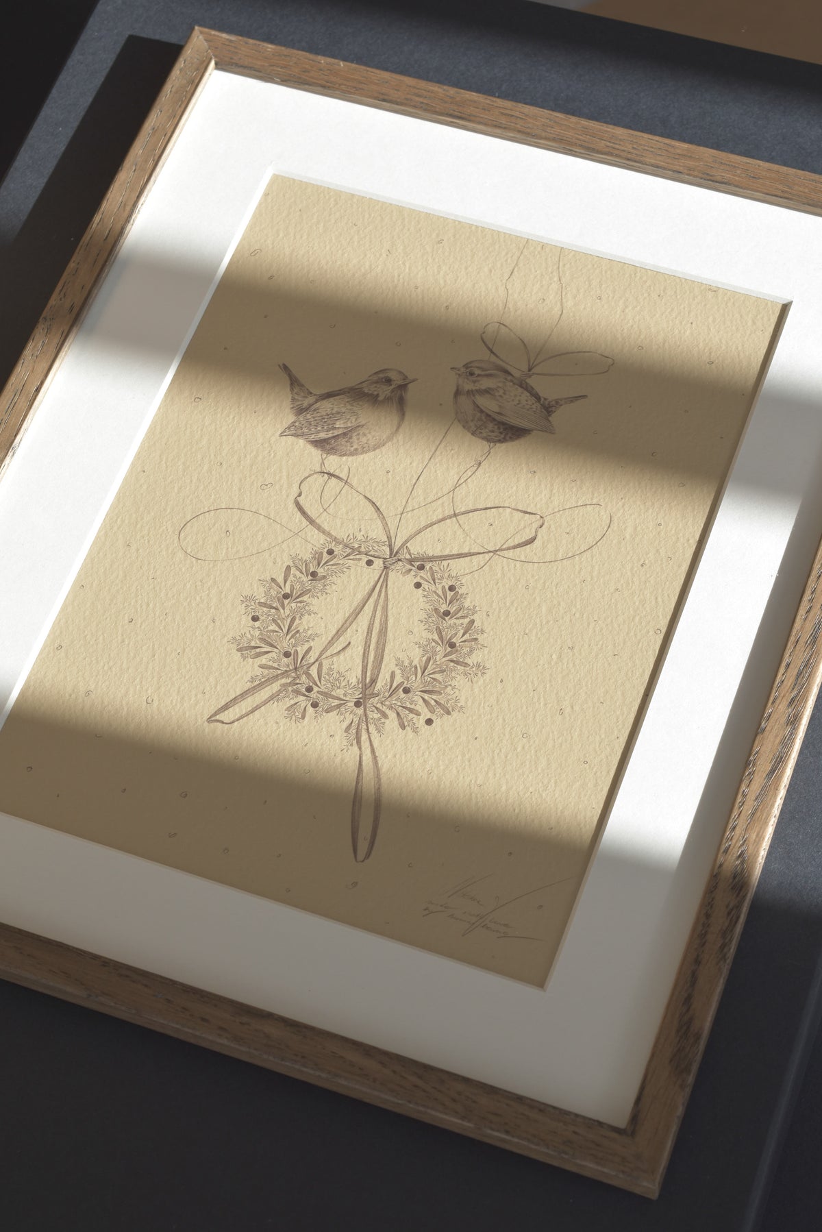 Framed art print featuring a delicate pencil sketch of two birds and a floral wreath on textured beige paper, set in a white mat and light wood frame with soft diagonal shadows.